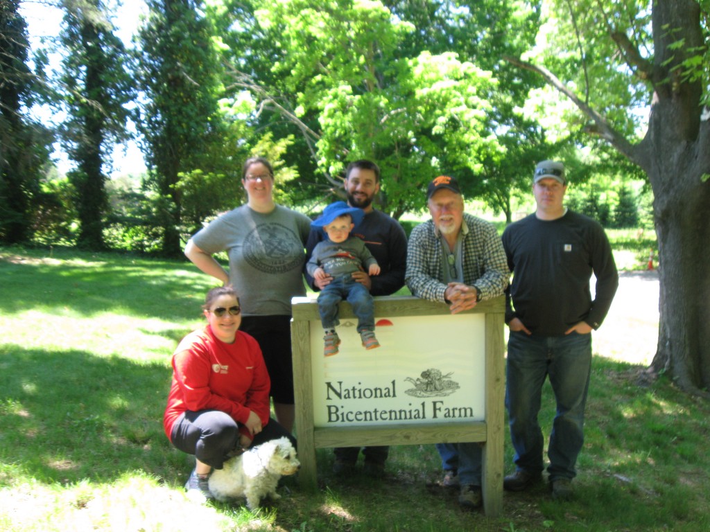 Rebecca, Abigail, Andrew, Bruce and Mike stand around the National Bicentennial Farm sign after a weekend of planting over 100 trees in Spring of 2015.  Logan sits on the sign's frame, and Lucy, a miniature schnoodle dog, sits in front of Rebecca.