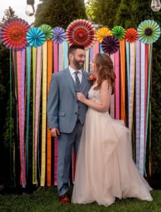 Andrew and Katie stand together at their wedding before a multicolored backdrop. Andrew is in a grey suit and Katie is in a blush wedding gown. They are looking at each other and smiling.