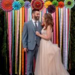 Andrew and Katie stand together at their wedding before a multicolored backdrop. Andrew is in a grey suit and Katie is in a blush wedding gown. They are looking at each other and smiling.